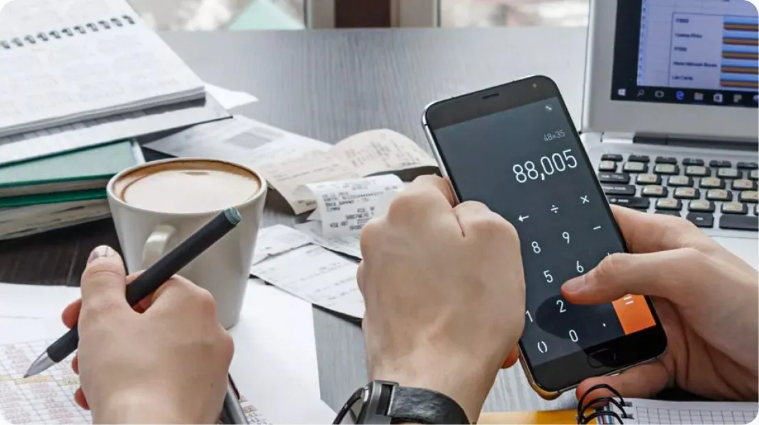 Hands using a calculator app beside a laptop and coffee
